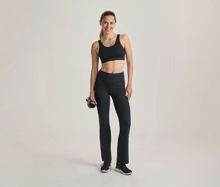 Lächelnde Frau in schwarzem Sport-BH und schwarzer Schlaghose, die eine schwarze Trinkflasche hält, steht vor hellem Hintergrund.