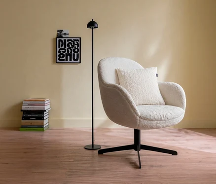 Ein offwhite Sessel mit einem offwhite SCHÖNER WOHNEN-Kollektion Dekokissenbezug »TEDDY« steht neben einer schwarzen Stehlampe. Links davon sind ein Bücherstapel und zwei Bilder an der hellgelben Wand zu sehen.