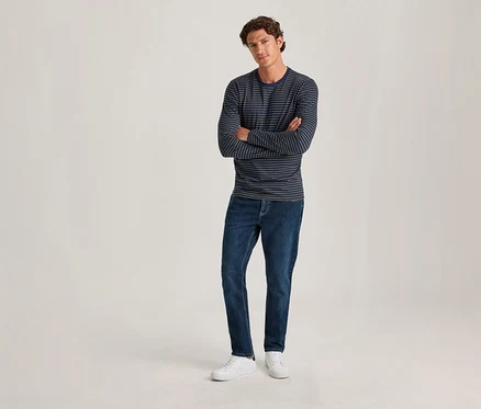 Ein Mann mit lockigem braunem Haar steht mit verschränkten Armen da. Er trägt ein dunkelblaues und hellblaues gestreiftes Langarmshirt, dunkelblaue Jeans und weiße Sneaker vor einem hellen, einfarbigen Hintergrund.