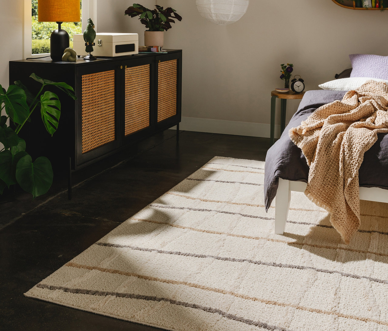 Ein Schlafzimmer mit einem hellen benuta Teppich #Junis# mit beigen und grauen Streifen. Links steht ein schwarzer Schrank mit Rattantüren, rechts ein Bett mit dunkelgrauer Bettwäsche und einer beigen Decke.