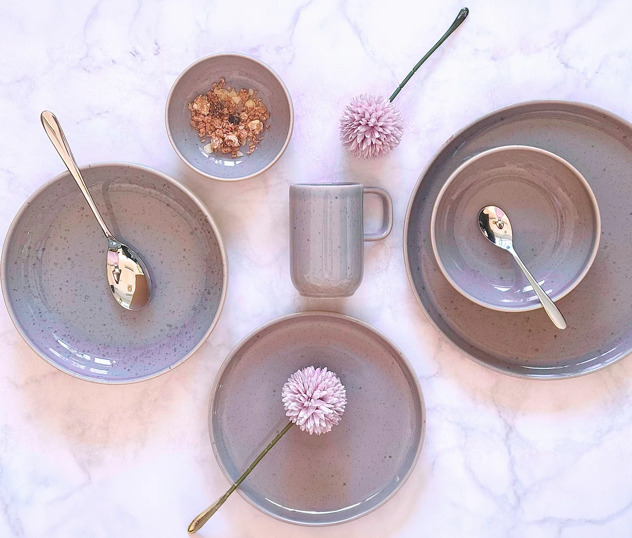 Ein hellgraues, gesprenkeltes Mäser Tafelservice »Candy« mit Tellern, Schalen und Tasse auf Marmor. Eine Schale enthält Müsli, zwei Löffel und zwei rosa Blumen sind ebenfalls zu sehen.
