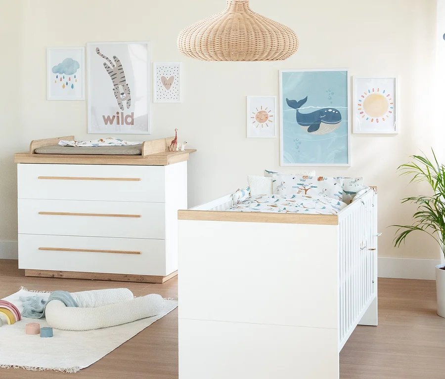 Ein Kinderzimmer mit einem weißen Roba Lumo 2-teiligen Kinderzimmer-Set, bestehend aus Wickelkommode und Babybett. Die Wickelkommode hat eine graue Auflage, das Bett Tier-Muster-Bettwäsche. An der Wand hängen Bilder von Tieren und Naturmotiven.