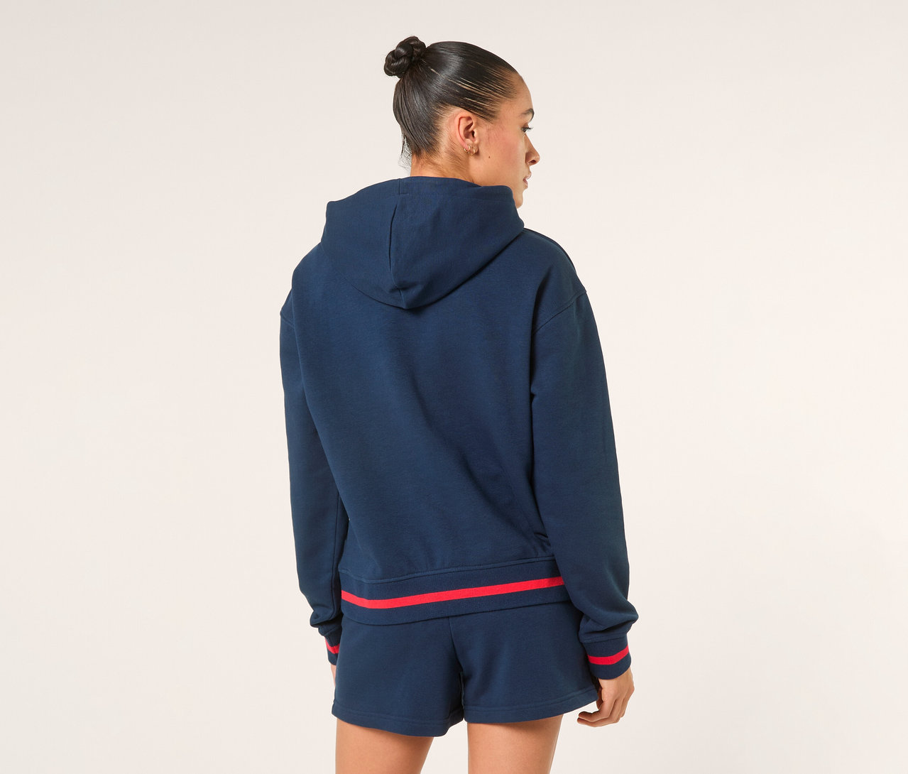 Frau von hinten, trägt einen dunkelblauen FILA Damen-Hoodie »MARLIA« mit roten Streifen an Ärmelbündchen und Saum sowie passende Shorts.