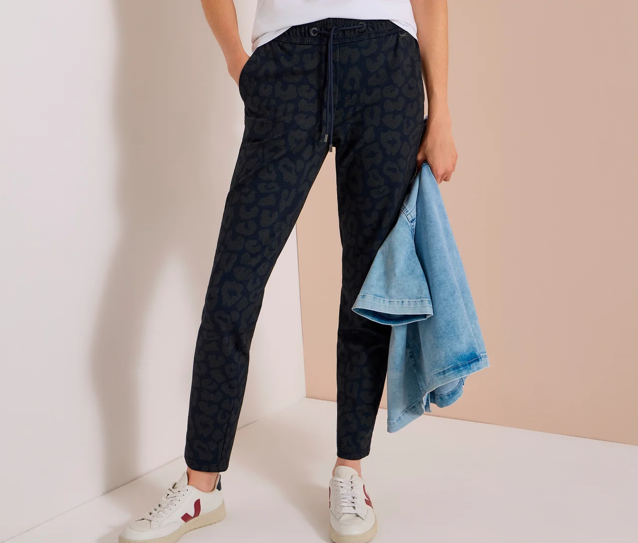 Eine Person trägt eine dunkelblaue CECIL Jerseyhose mit Alloverprint, ein weißes T-Shirt und weiße Sneaker. Sie hält eine hellblaue Jeansjacke.