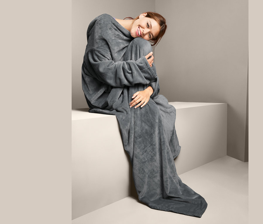 Frau sitzt auf einem weißen Block und ist in eine graue Kuscheldecke mit integrierten Ärmeln gehüllt.