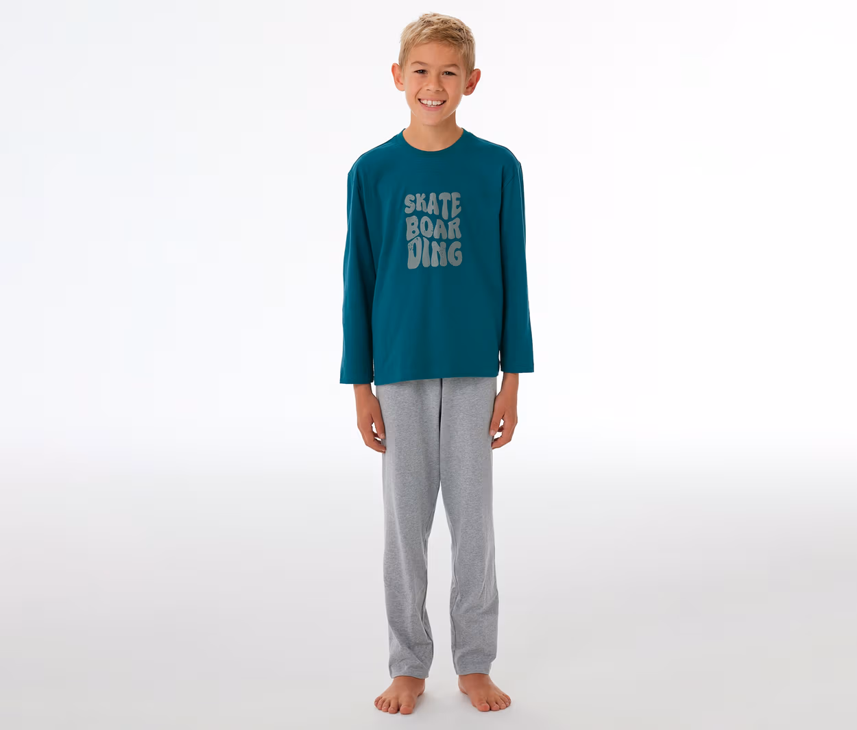 Lächelnder Junge in einem Kinder-Pyjama mit petrolfarbenem Langarmshirt, grauer "SKATE BOARDING"-Aufschrift und grauer Pyjamahose.