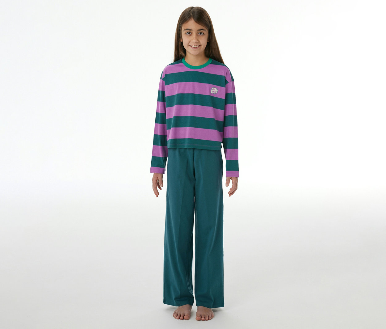 Mädchen lächelt, trägt einen SCHIESSER Kinder-Pyjama mit lila-grün gestreiftem Langarmshirt, grüner Hose und hat nackte Füße.