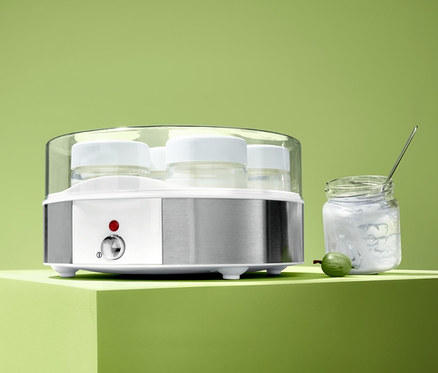 Elektrischer Joghurtbereiter mit sieben Gläsern steht neben einem Glas Joghurt und einer grünen Stachelbeere.
