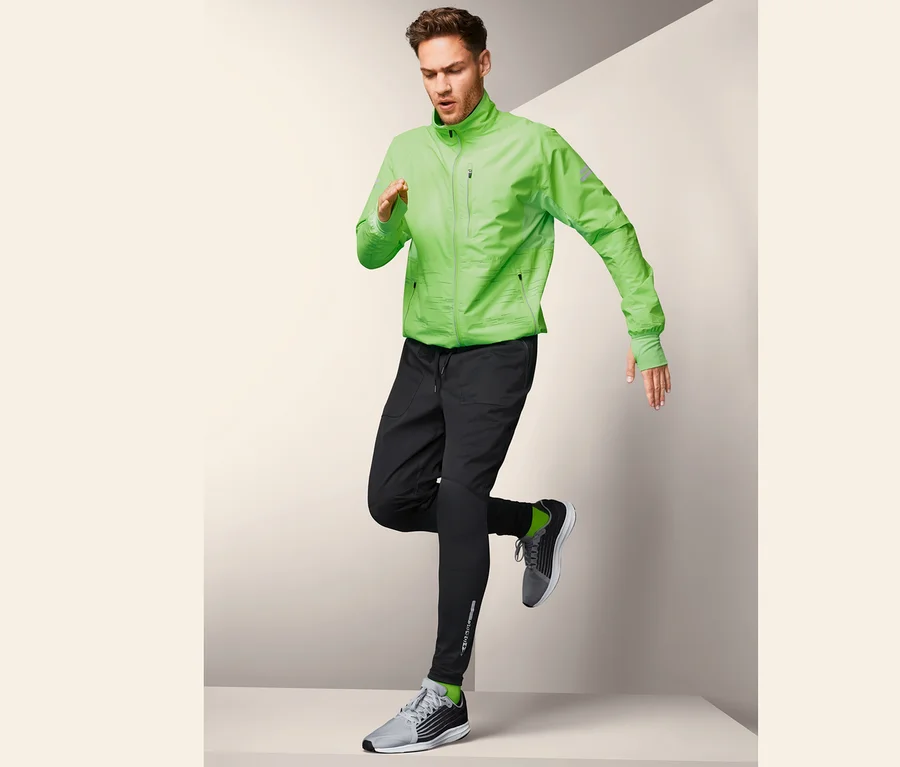 Mann in grüner Jacke und schwarzer Windprotection-Sporttight posiert beim Laufen.