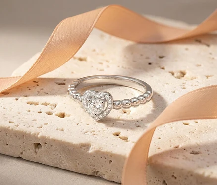 Silberner Amor Ring mit Herz-Symbol, besetzt mit Diamanten, liegt auf einem hellen Stein neben einem rosafarbenen Band.