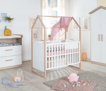 Babyzimmer mit Wickelkommode aus hellem Holz und Weiß, einem weißen Gitterbett mit Hausrahmen und rosa Decke, sowie einem passenden weißen Kleiderschrank mit Dachform.
