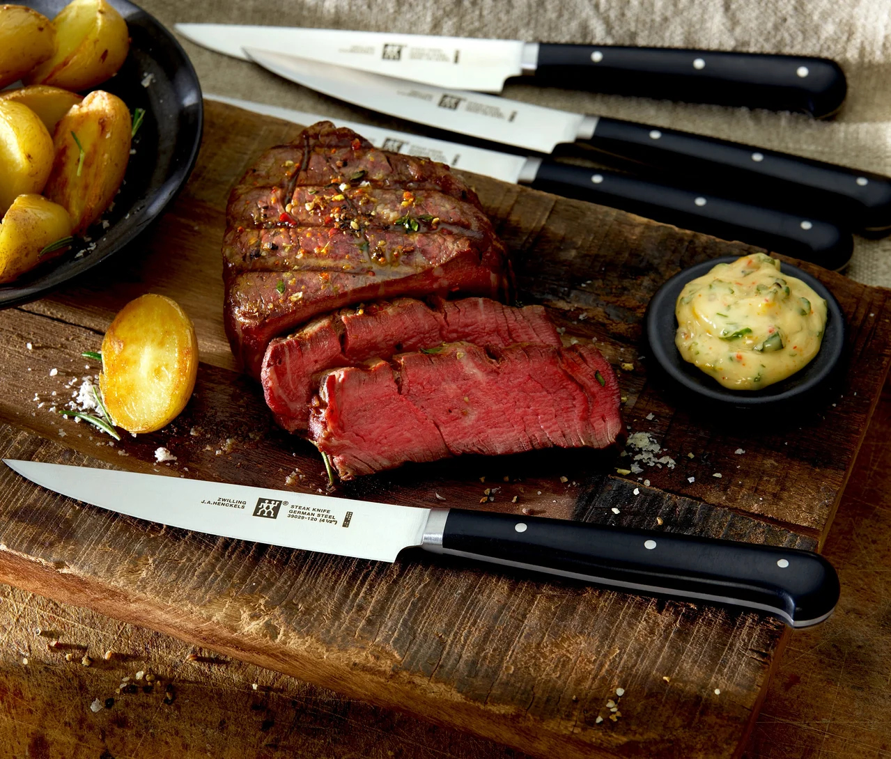 Gegrilltes Steak, teilweise geschnitten, mit Bratkartoffeln, einer Sauce und vier ZWILLING Steakmessern aus dem 4-teiligen Set auf einem Holzbrett.