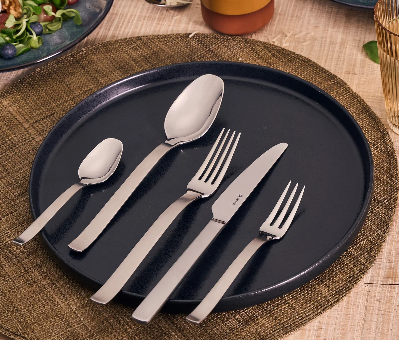 Ein Henckels by ZWILLING "Clouds" Besteckset mit großem Löffel, kleinem Löffel, Gabel, Messer und Dessertgabel auf einem dunkelblauen Teller. Das silberne Besteck hat matte Griffe und polierte Köpfe. Der Teller steht auf einem geflochtenen Platzset, mit Salatteller und Glas im unscharfen Hintergrund.
