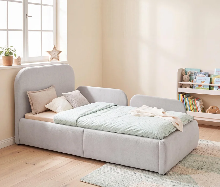 Ein hellgraues, gepolstertes Kinderbett mit abgerundetem Kopfteil und Seitenteilen, bedeckt mit einem hellgrün gemusterten Bettbezug und mehreren Kissen. Das Bett steht auf einem hellen Holzfußboden mit einem gemusterten Teppich. Hinter dem Bett befindet sich ein Fenster mit einer Pflanze und einer Sternendekoration, rechts davon ein kleines weißes Bücherregal mit Büchern und Spielzeug.