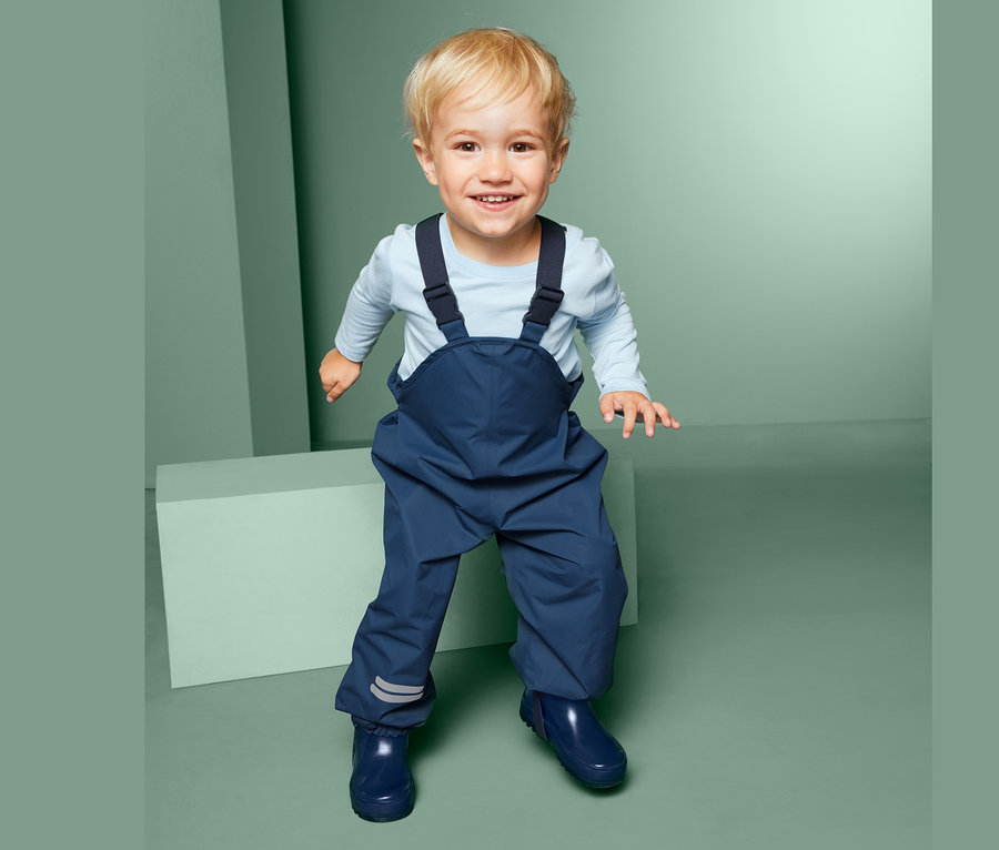 Ein lächelndes Kleinkind steht und trägt eine blaue Regenhose mit Latz und blaue Gummistiefel.