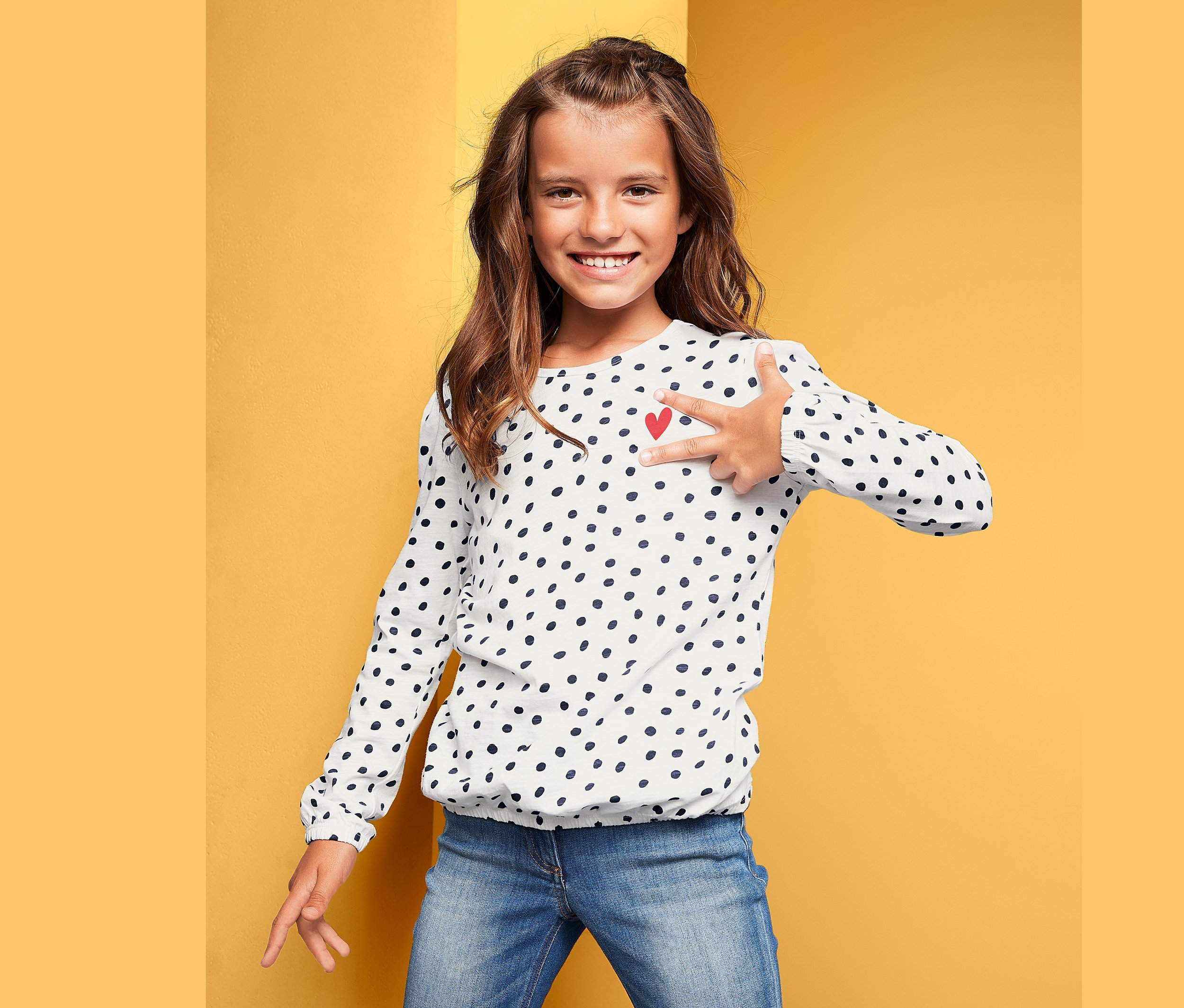 Lächelndes Mädchen zeigt auf Langarmshirt mit Punktmuster und rotem Herz.