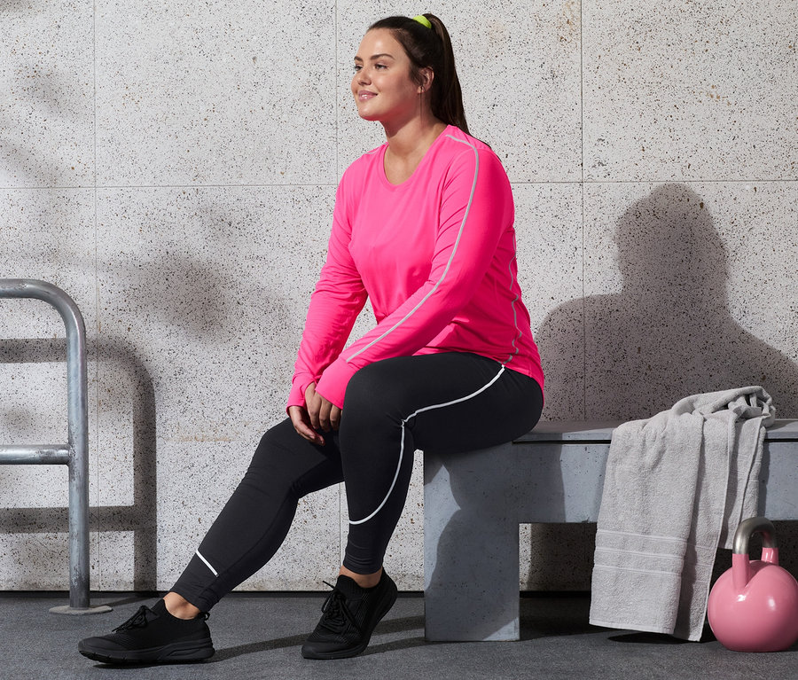 Frau sitzt in neonpinkem Langarm-Funktionsshirt und Sporttight auf einer Bank.
