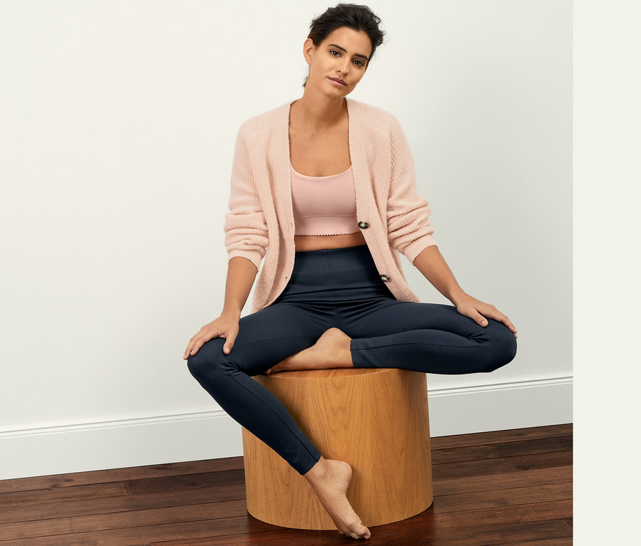 Eine Frau sitzt auf einem Holzhocker und trägt Leggings aus Heavy-Jersey, ein rosa Tanktop und eine rosa Strickjacke.