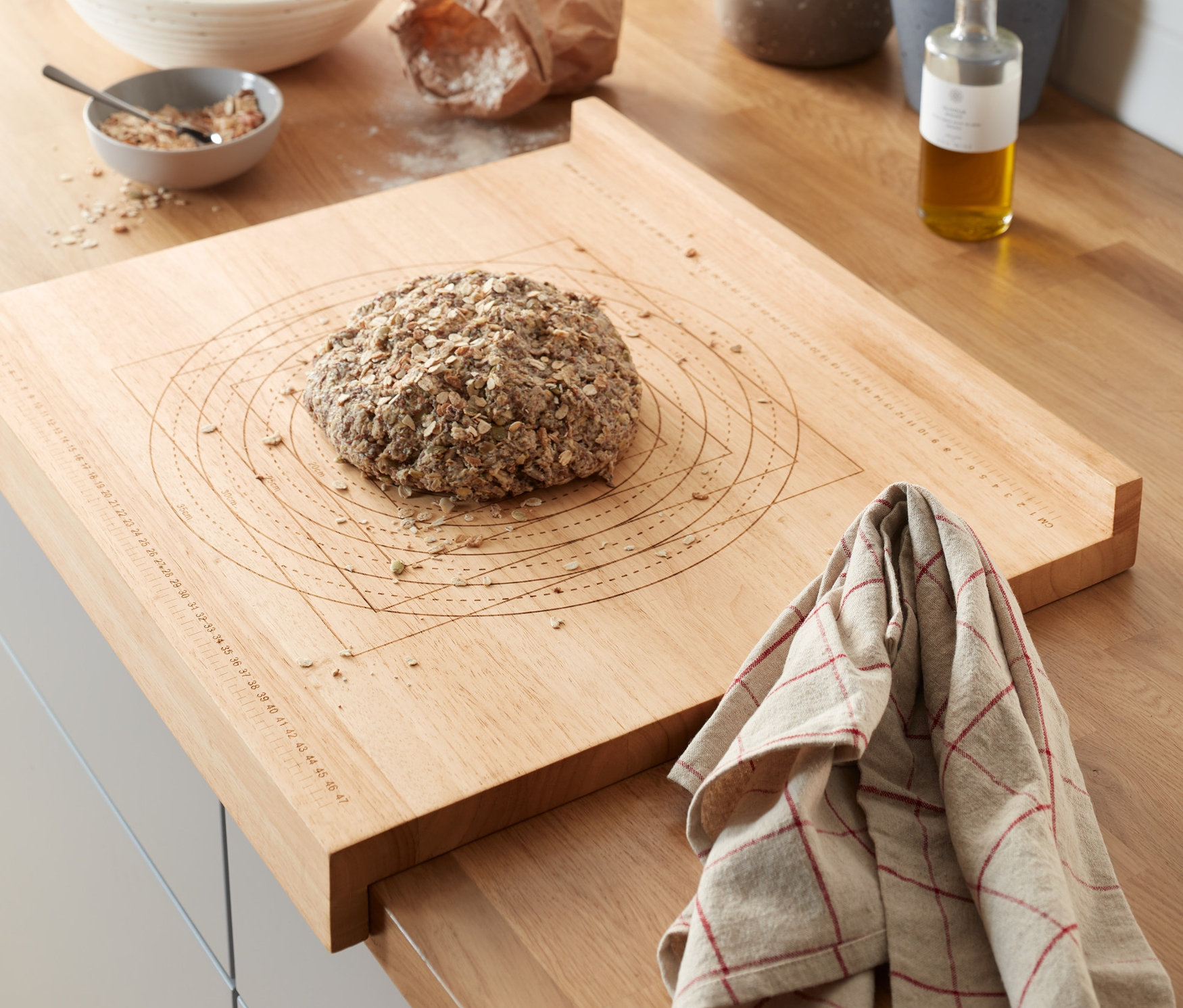 Auf einer Küchentheke liegt ein hölzernes Back- und Arbeitsbrett mit einem Brotlaib. Im Hintergrund sind Kräuter in Töpfen und andere Küchenutensilien zu sehen.