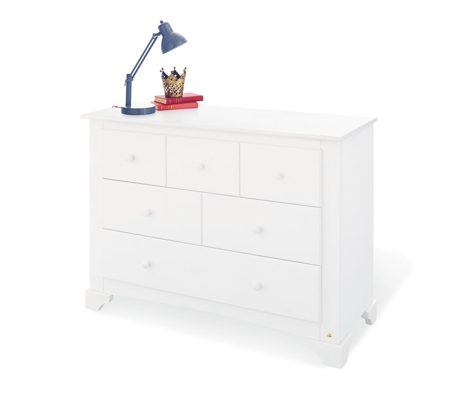 Weiße Kommode mit sechs Schubladen, auf der ein Buch, eine Krone und eine Lampe liegen.