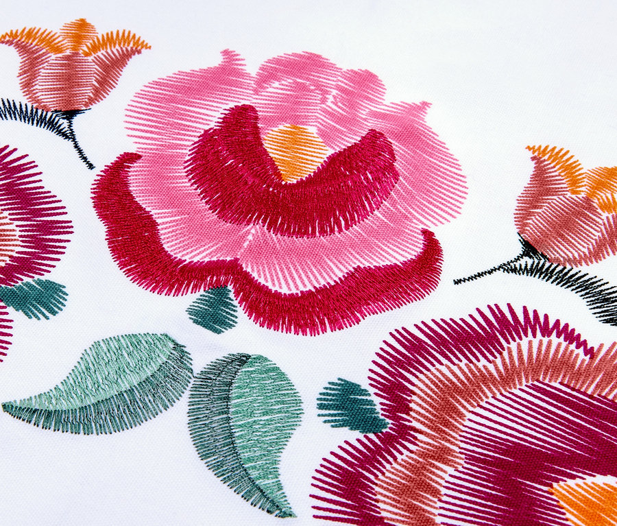 Nahaufnahme eines Tischläufers mit Blumenstickerei in Rosa-, Rot- und Grüntönen auf weißem Hintergrund.