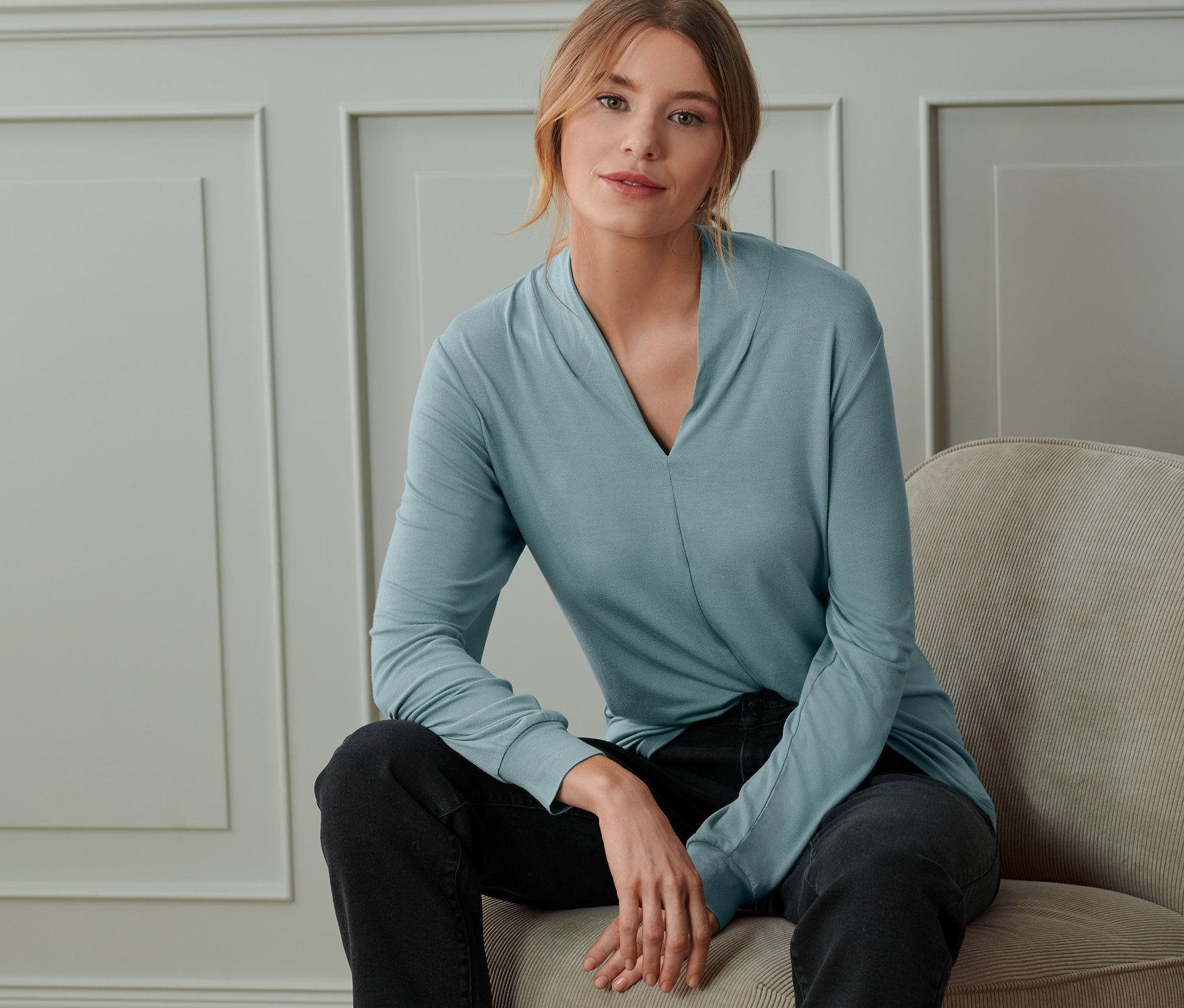 Eine Frau sitzt auf einem Stuhl und trägt ein rauchblaues Langarmshirt mit V-Ausschnitt.