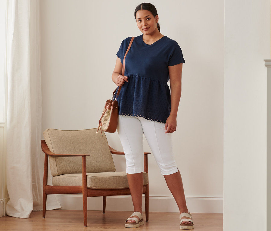 Eine Frau steht und trägt ein dunkelblaues Shirt im Materialmix mit Stickerei, eine 3/4-Stretchhose, Plateau-Sandalen aus Leder und eine Beuteltasche.