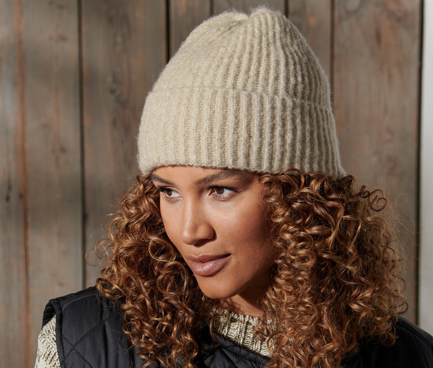Porträt einer Frau mit braunen lockigen Haaren, die eine Strickmütze und eine schwarze Weste trägt.