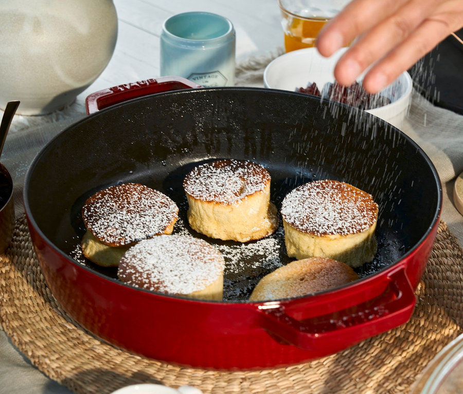 Eine Hand streut Puderzucker auf Pfannkuchen in einem roten STAUB Gusseisen-Bräter »Braisers« mit Glasdeckel, 28 cm.