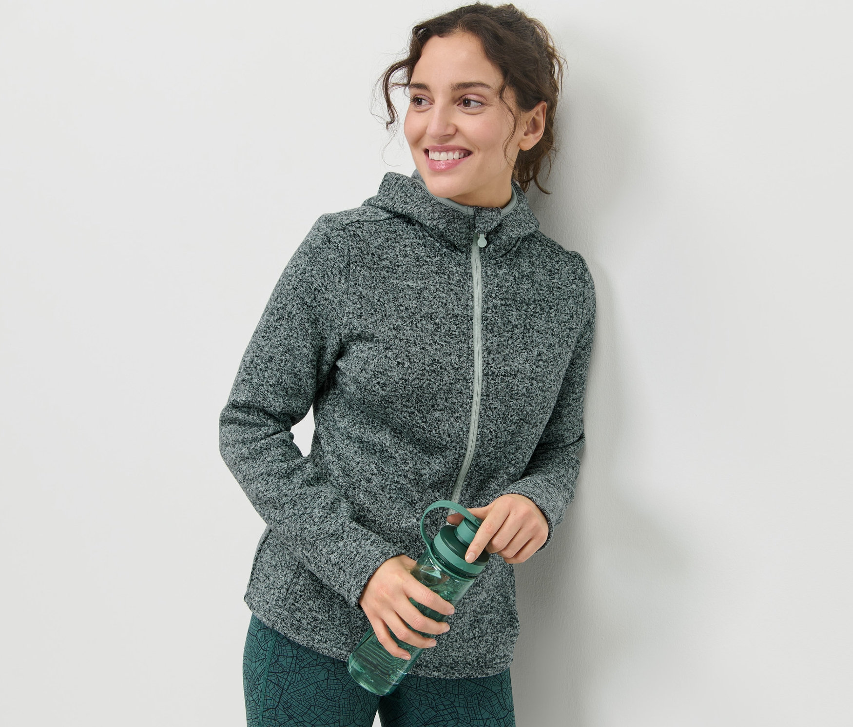 Frau lächelt und hält eine grüne Strickfleece-Jacke und eine Wasserflasche.