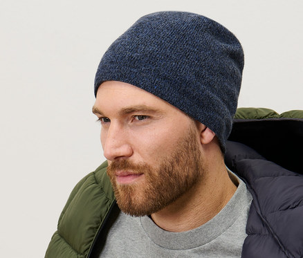 Mann trägt eine dunkelblau melierte Strickmütze, einen grauen Pullover und eine grüne Daunenjacke.