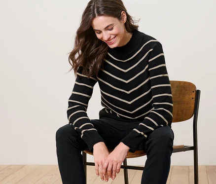 Frau sitzt auf einem Stuhl und trägt einen gestreiften Strickpullover mit Stehkragen, schwarz.