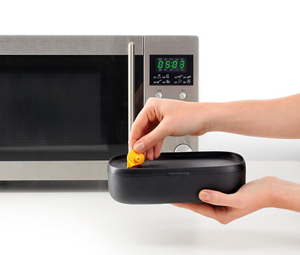 Eine Frau legt einen gelben Gegenstand in ein schwarzes Lunch-Box-Set mit Besteck neben einer Mikrowelle.