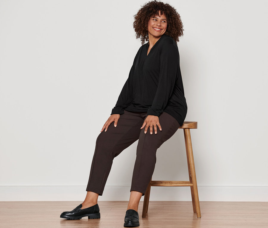 Frau sitzt auf einem Hocker und trägt ein schwarzes Langarmshirt mit V-Ausschnitt, eine dunkelbraune Joggpants mit Biese und Leder-Loafer.