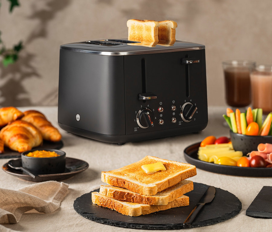 Auf einem Tisch stehen ein schwarzer WILFA Toaster FAMILY mit Toast, Croissants, Marmelade, Gemüse und Getränke.