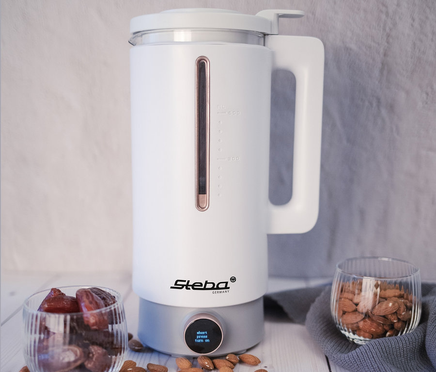 Weißer Steba Vegan-Drink-Maker VDM 2 HOT & COLD steht auf einem Holztisch neben Gläsern mit Datteln und Mandeln.