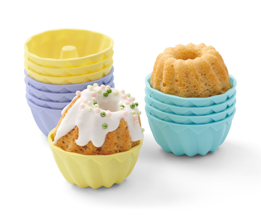 Mini-Gugelhupfe in bunten Förmchen. Ein Gugelhupf ist mit Zuckerguss und bunten Streuseln verziert. Im Hintergrund stehen 12 Mini-Gugelhupfformen.