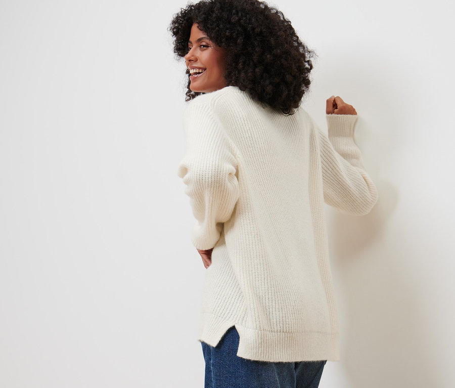Frau blickt über die Schulter und posiert in einem Strickpullover mit Herz-Jacquard und Jeans.