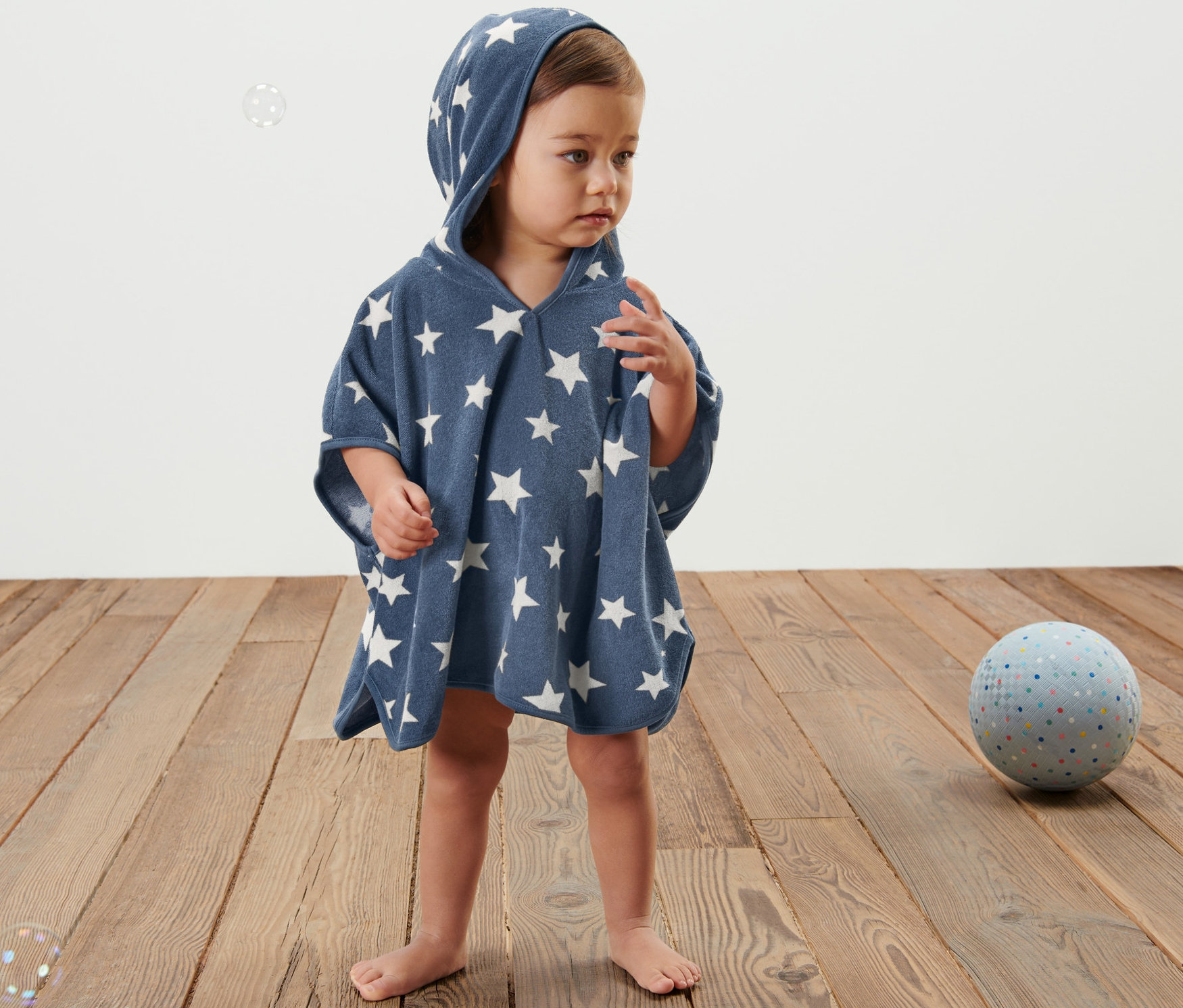 Ein Kind steht auf einem Holzboden und trägt einen blauen Frottierponcho mit Kapuze und weißen Sternen.