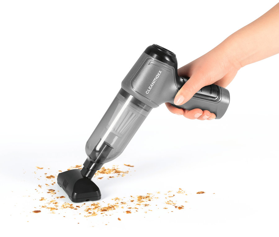 Eine Hand hält einen grauen Akku-Handstaubsauger und saugt Krümel auf einer weißen Oberfläche auf.
