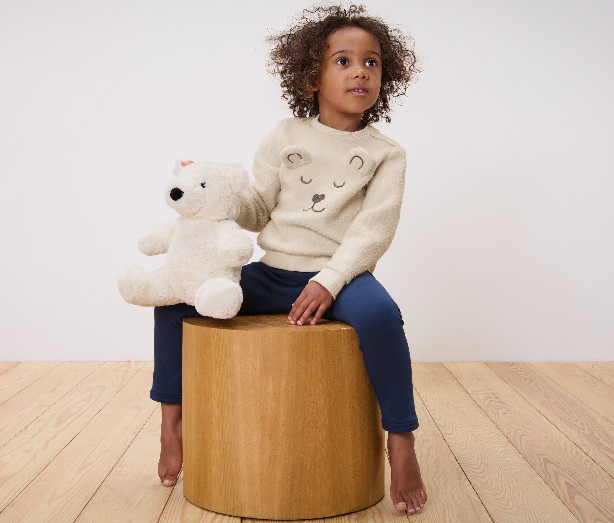 Mädchen sitzt auf einem Holzhocker und hält einen Teddybären. Sie trägt einen Kinder-Teddy-Sweater.