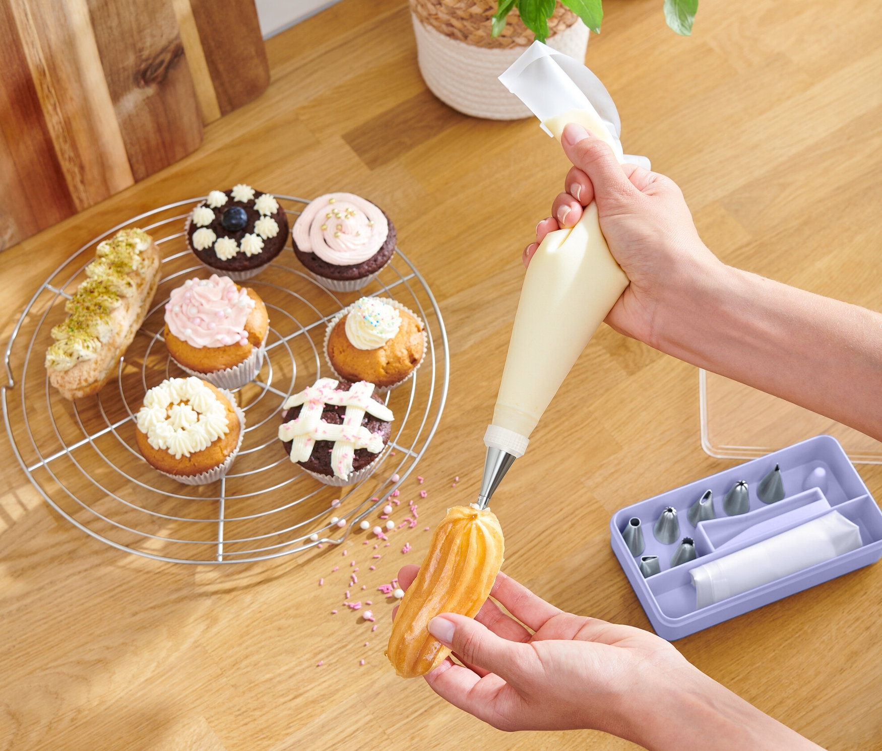 Eine Person dekoriert ein Eclair mit Creme, wobei sie ein Spritztüllen-Set verwendet. Auf dem Tisch liegen Muffins und eine Schale mit Früchten.