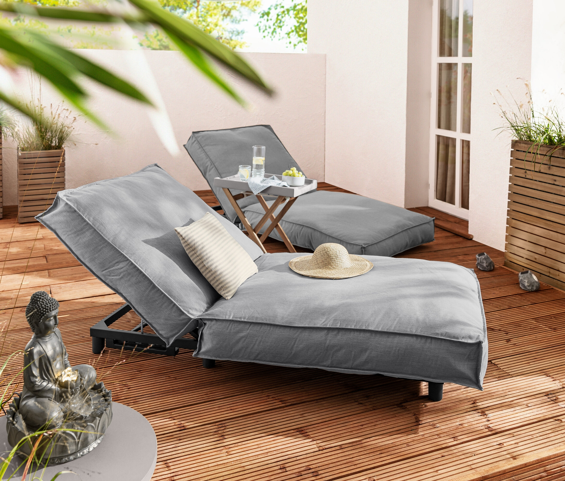 Zwei graue Liegen mit Kissen und Hut, Tisch mit Getränken, 3 Solar-LED-Steine und Buddha-Gartenbrunnen auf einer Holzterrasse.