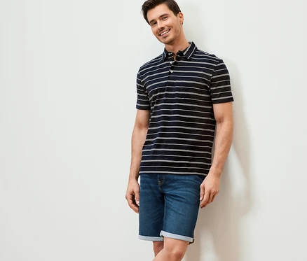 Mann lächelt an eine Wand gelehnt. Er trägt ein dunkelblau gestreiftes Jersey-Poloshirt und Jeansshorts.