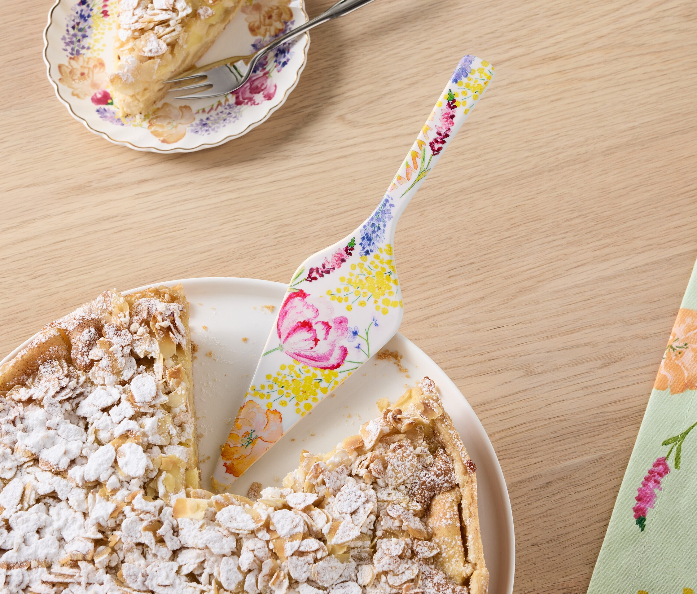 Ein angeschnittener Mandelkuchen auf einem Teller mit Tortenheber mit Blumenmuster, daneben ein Stück Kuchen auf einem Teller.
