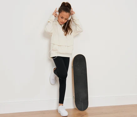Ein Mädchen mit braunen, hochgesteckten Haaren hört Musik mit Kopfhörern, lehnt an einer Wand neben einem Skateboard. Sie trägt einen Pullover und 2 Kinder-Leggings.
