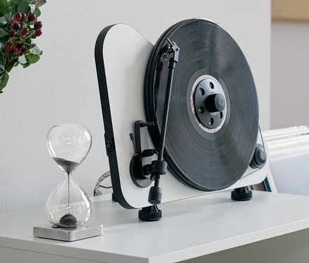 Auf einer weißen Kommode stehen eine magnetische Sanduhr, ein Stereo Regallautsprecher mit Bluetooth® und ein Vertikaler Plug-and-Play-Plattenspieler »Pro-Ject VT-E BT« mit Bluetooth®.