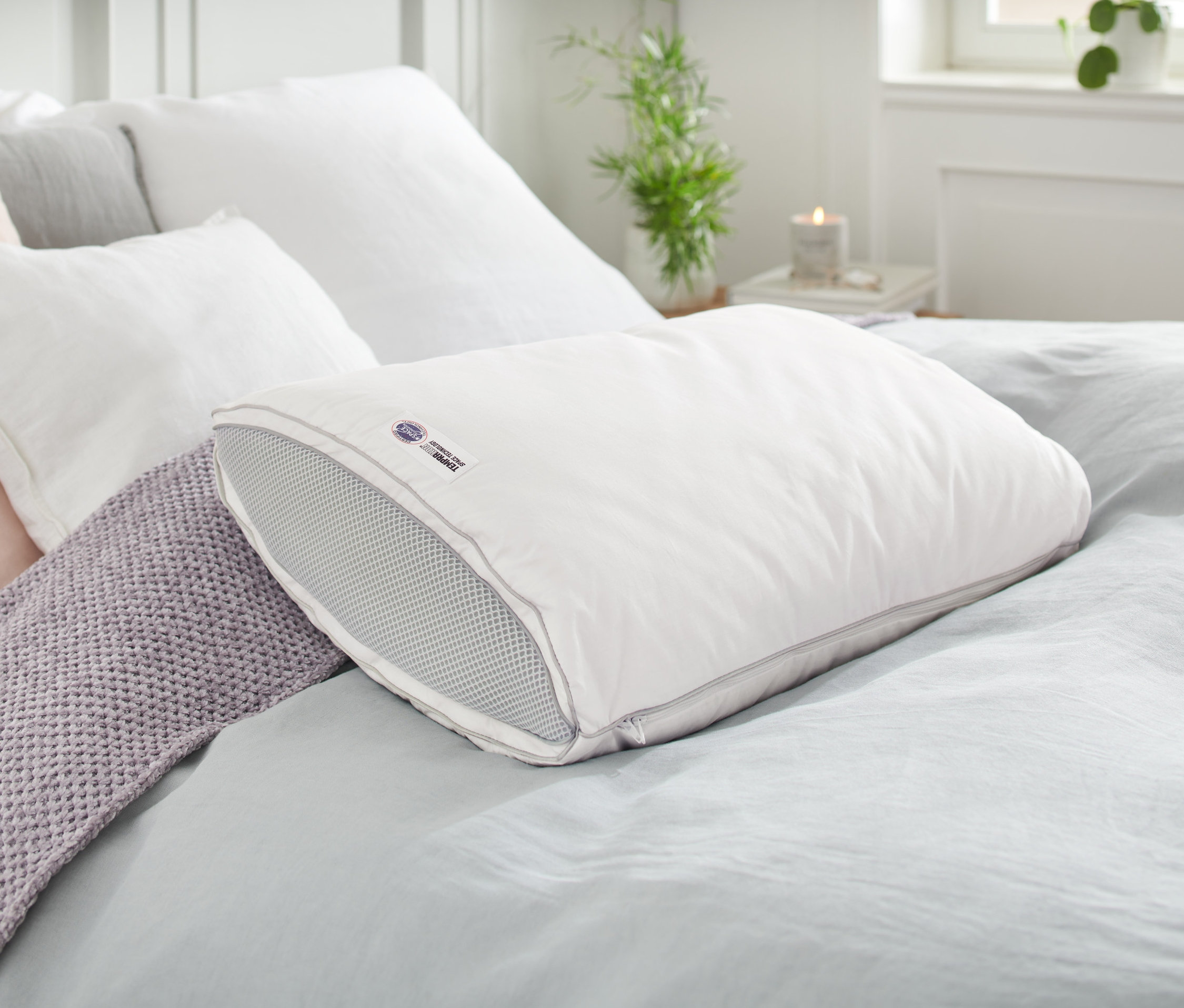 Ein Temprakon V-Zone Kissen liegt auf einem Bett mit anderen Kissen und einer grauen Decke.