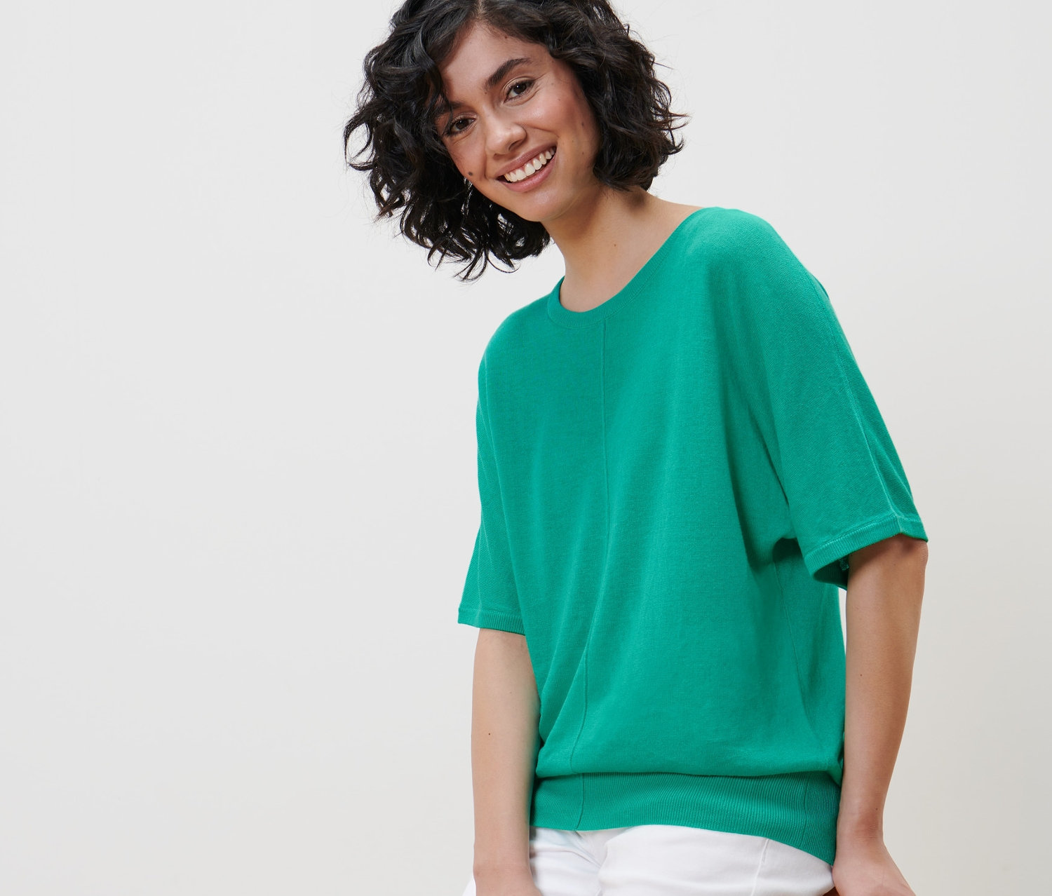 Frau sitzt auf einem Stuhl und trägt ein grünes Feinstrick-Shirt mit Leinen und eine weiße Hose.