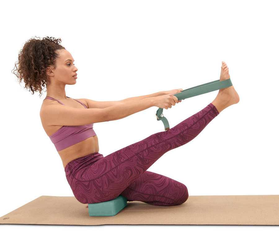 Frau sitzt mit Yoga-Block auf einer Yoga-Matte und benutzt einen Yoga-Gurt aus dem Yoga-Set.